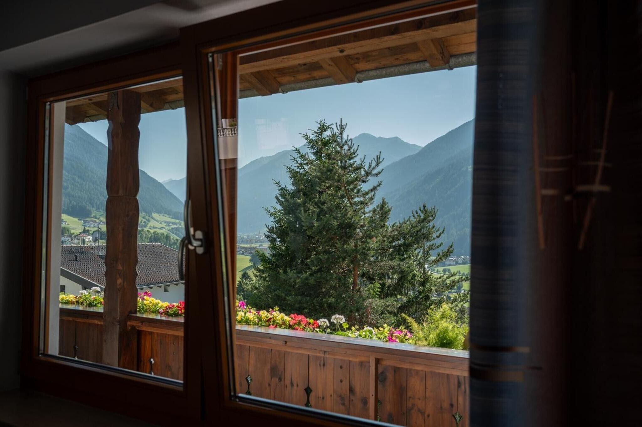 Ein Fenster mit Holzrahmen, das einen Blick auf eine gebirgige Landschaft und eine Holzterrasse mit bunten Blumen bietet.