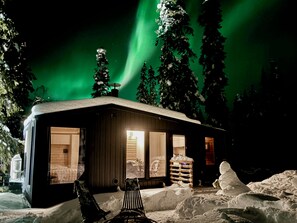 Una cabaña con fachada de madera, un dormitorio visible con cama y una silla, y suelo cubierto de nieve.