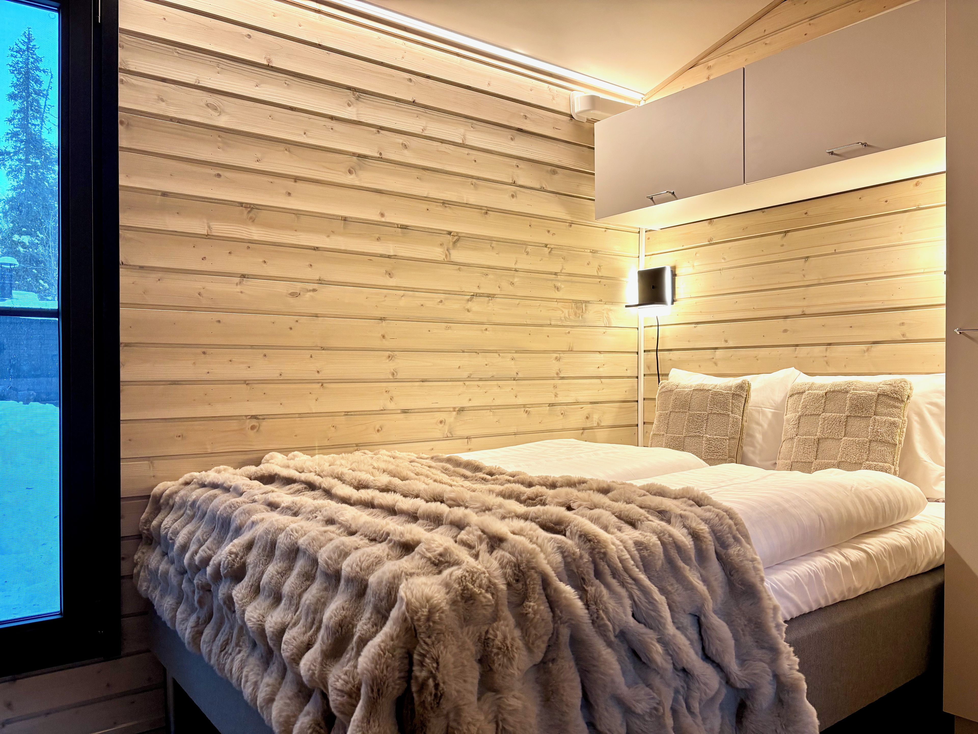 Een slaapkamer met een wand van houten latten, een bed met witte beddengoed en een raam met uitzicht op bomen.