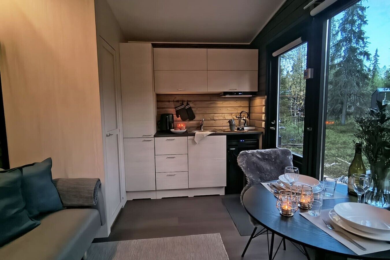 A compact kitchen with white cabinets, a dining table set for two, and a view of the outdoors through a large window.