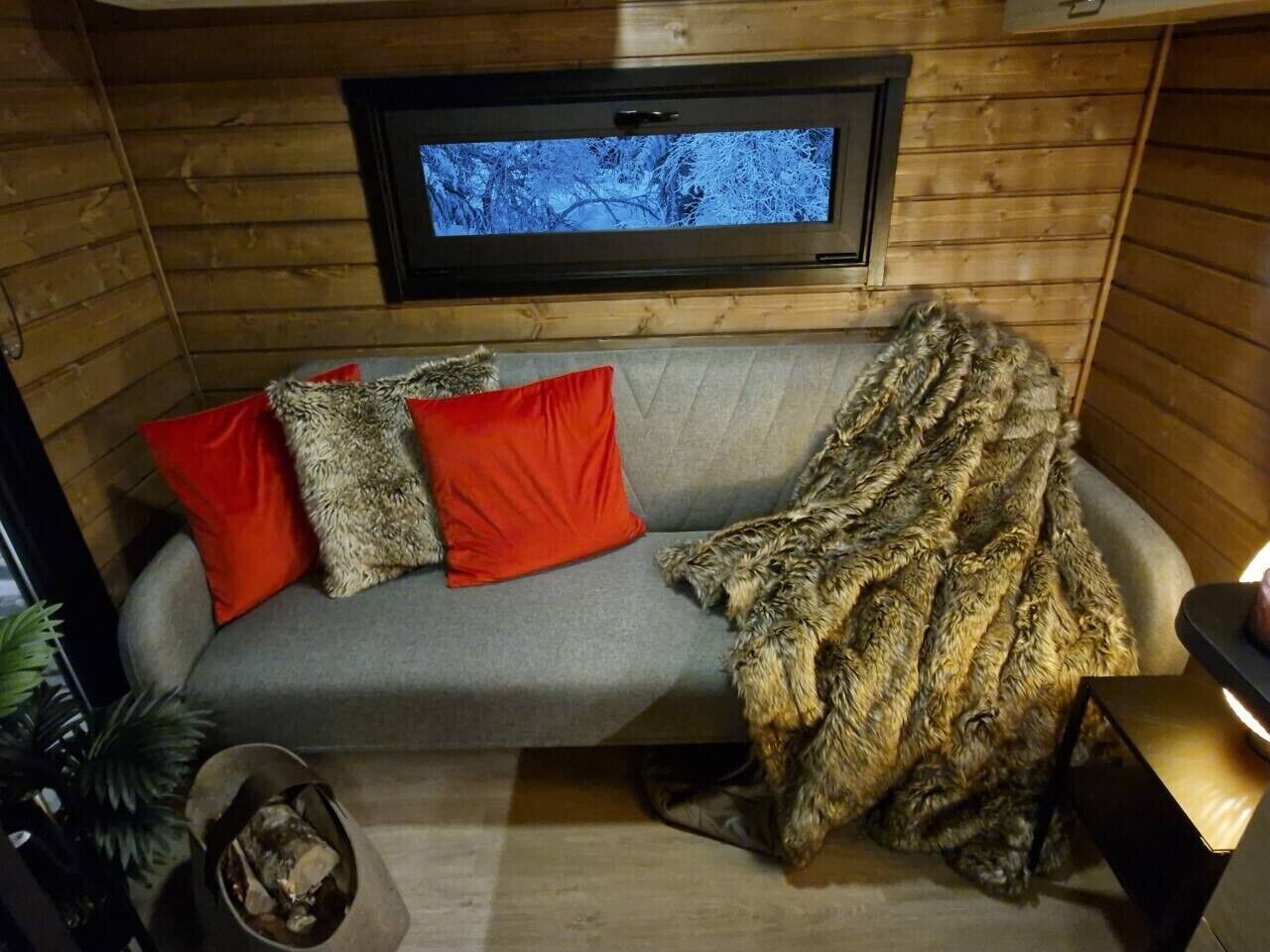 A cozy cabin interior with a grey sofa, red and brown cushions, a television mounted on the wall, and a window with a view of trees.