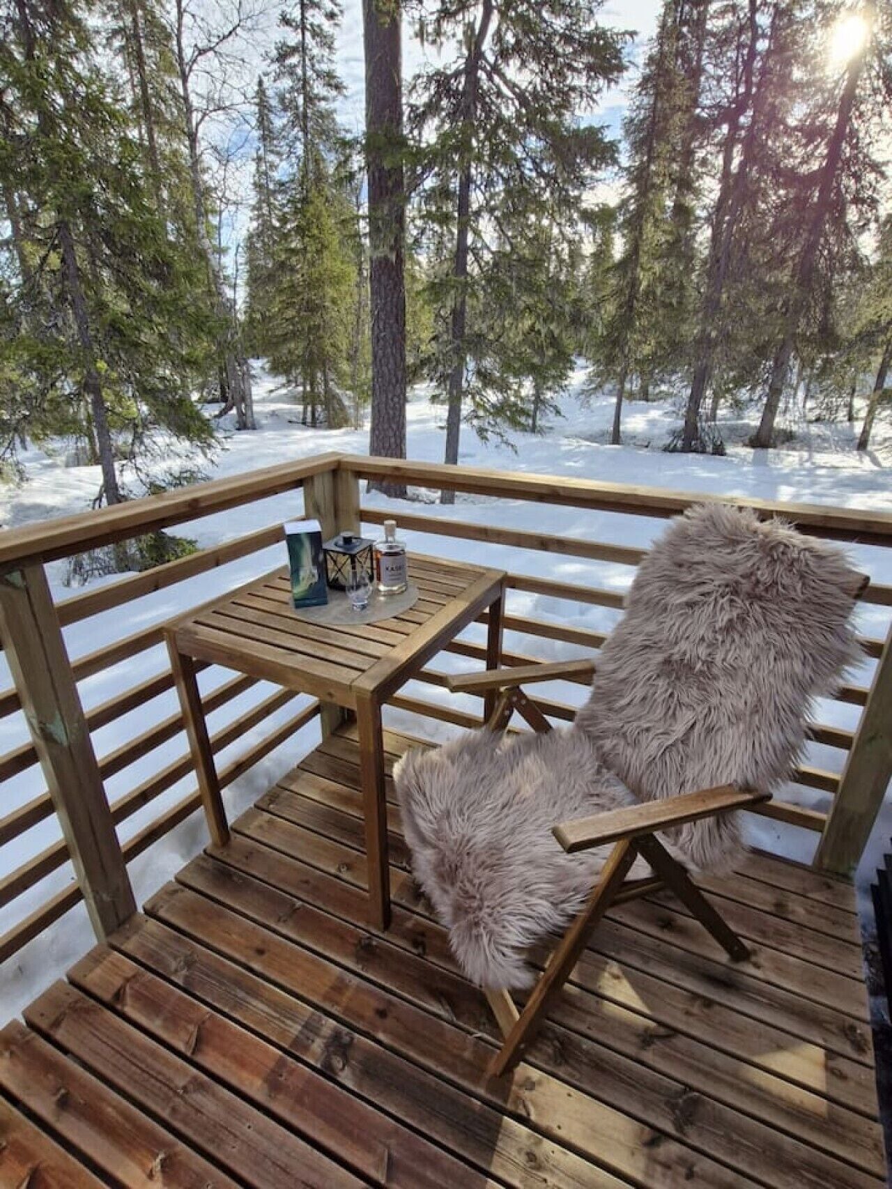 A wooden deck with a table and a chair, surrounded by snow-covered ground and trees.