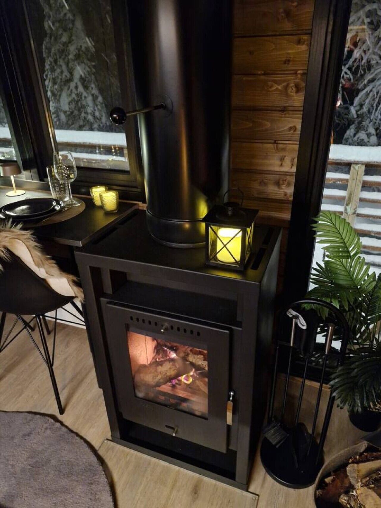 En vedeldad kamin med en glaslucka, ett litet bord med ett ljus och en stol.
