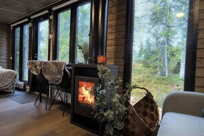 Una habitación acogedora con estufa de leña, ventanas amplias y vista al bosque.