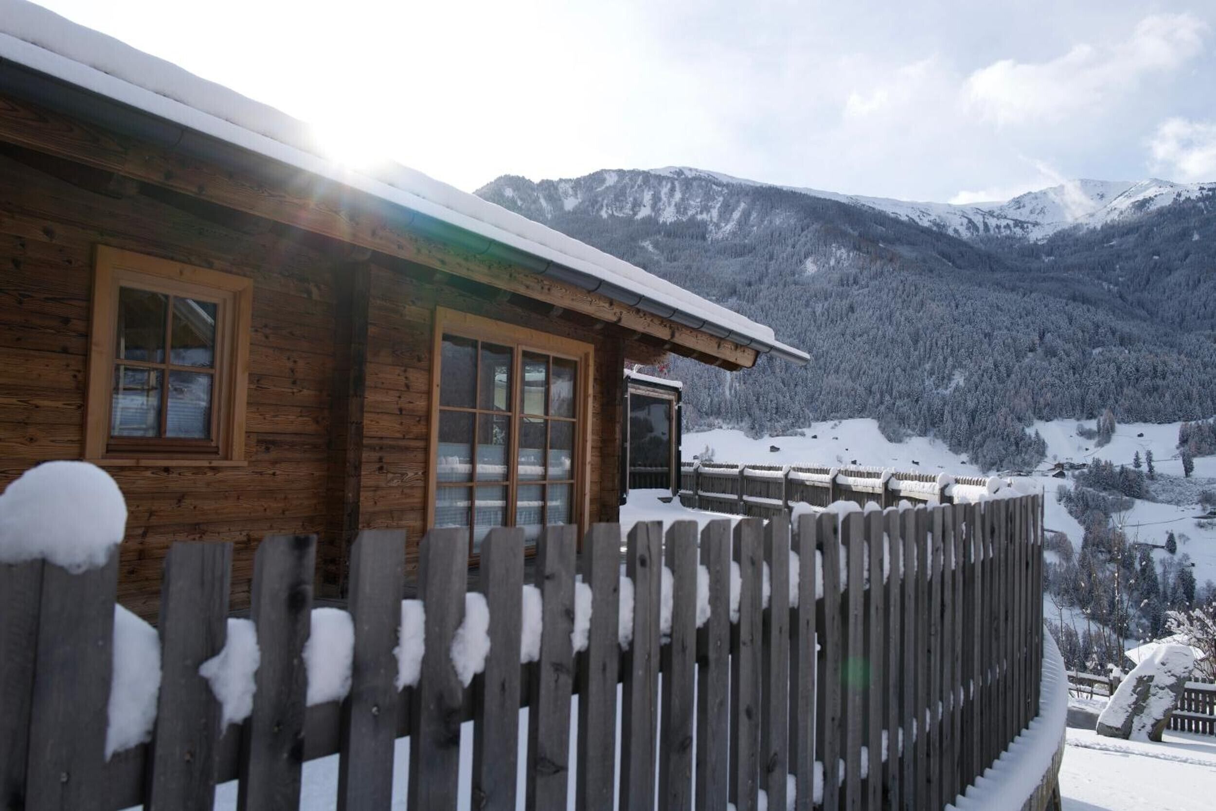 Uma cabana de madeira com grandes janelas, varanda e vista para montanhas cobertas de neve.