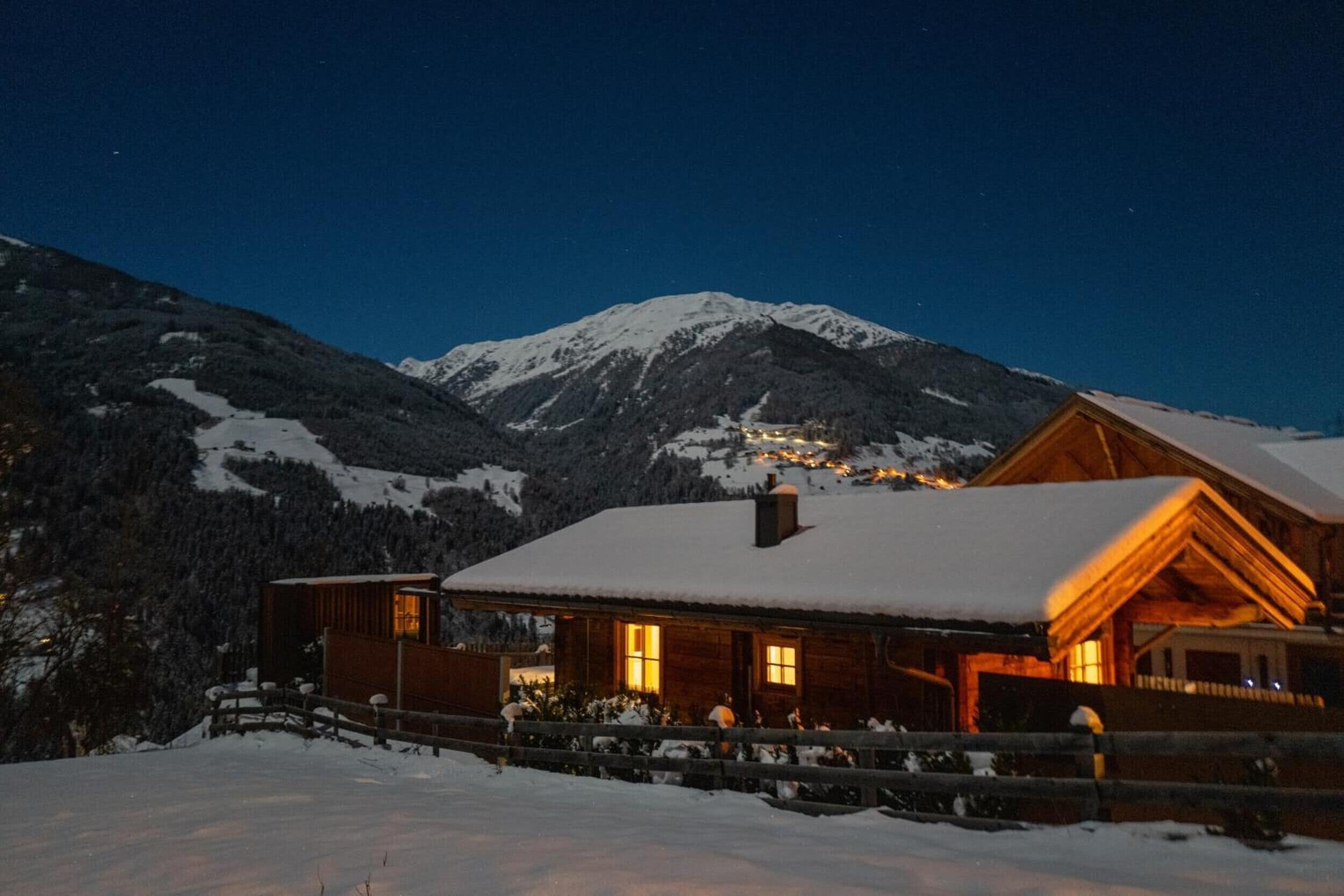 Eine Holzhütte mit schneebedecktem Dach, vor einer Kulisse schneebedeckter Berge und eines klaren Nachthimmels.