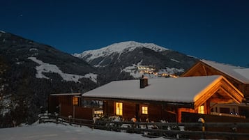 Eine Holzhütte mit schneebedecktem Dach, vor einer Kulisse schneebedeckter Berge und eines klaren Nachthimmels.