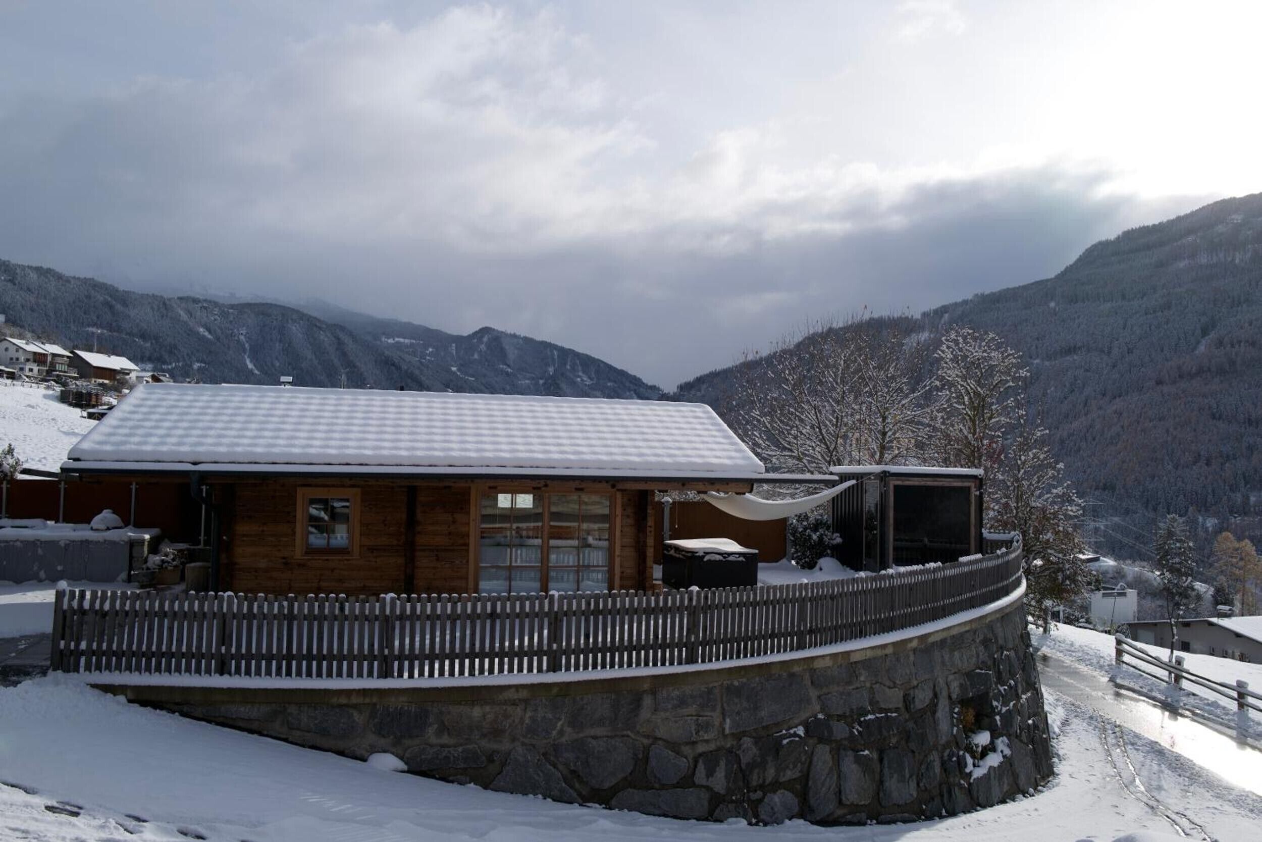 Een houten chalet met een stenen steunmuur, omgeven door met sneeuw bedekte bergen.