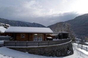 Una cabaña de madera con un muro de contención de piedra, rodeada de montañas nevadas.