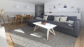 Uma sala de estar moderna com um sofá cinzento, uma mesa de centro branca e um espaço de jantar com cadeiras de madeira.