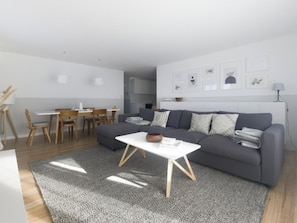 Ein modernes Wohnzimmer mit grauem Sofa, weißem Couchtisch und einem Essbereich mit Holzstühlen.