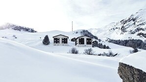Paisagem coberta de neve com dois edifícios, um maior e outro menor, rodeados por montanhas e um céu limpo.