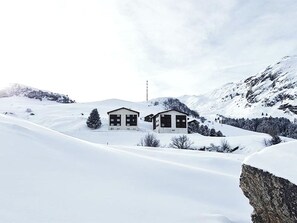 Schneebedeckte Landschaft mit zwei Gebäuden, einem größeren und einem kleineren, umgeben von Bergen und einem klaren Himmel.
