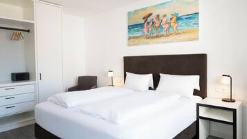 Ein ordentlich bezogenes Bett mit weißen Leinentüchern, ein dunkles Kopfteil und Nachttischlampen. Ein weißer Kleiderschrank mit Kleiderstange befindet sich links, und ein Gemälde mit Strandmotiv hängt über dem Bett.