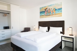 A neatly made bed with white linens, a dark headboard, and bedside lamps. A white wardrobe with a hanging rack is to the left, and a painting of a beach scene hangs above the bed. - Deluxe Apartment (Scharbeutz)