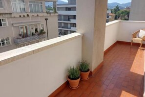 Un balcó amb rajoles de terracota, dues plantes en testos i una cadira de vimet.