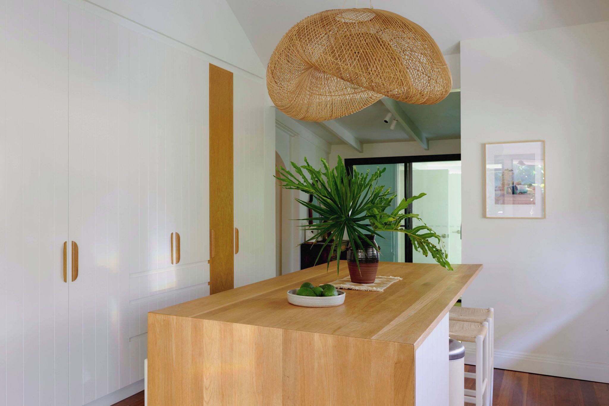 A kitchen with a wooden island, white cabinets, a woven pendant light, a potted plant, and a framed picture on the wall.