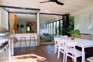 An open-plan living space with a dining area, bar stools, and a view through large windows. - Bruns Surf Shack: Beachy Hideaway. Pet friendly! (Brunswick Heads)