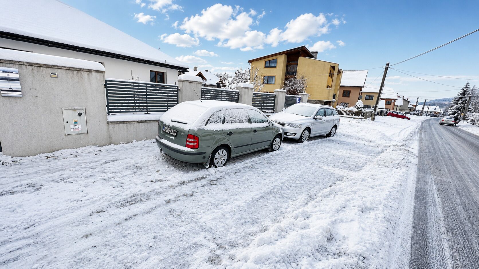 A residential street with two cars parked on the side, a building with a flat roof, and a clear sky.