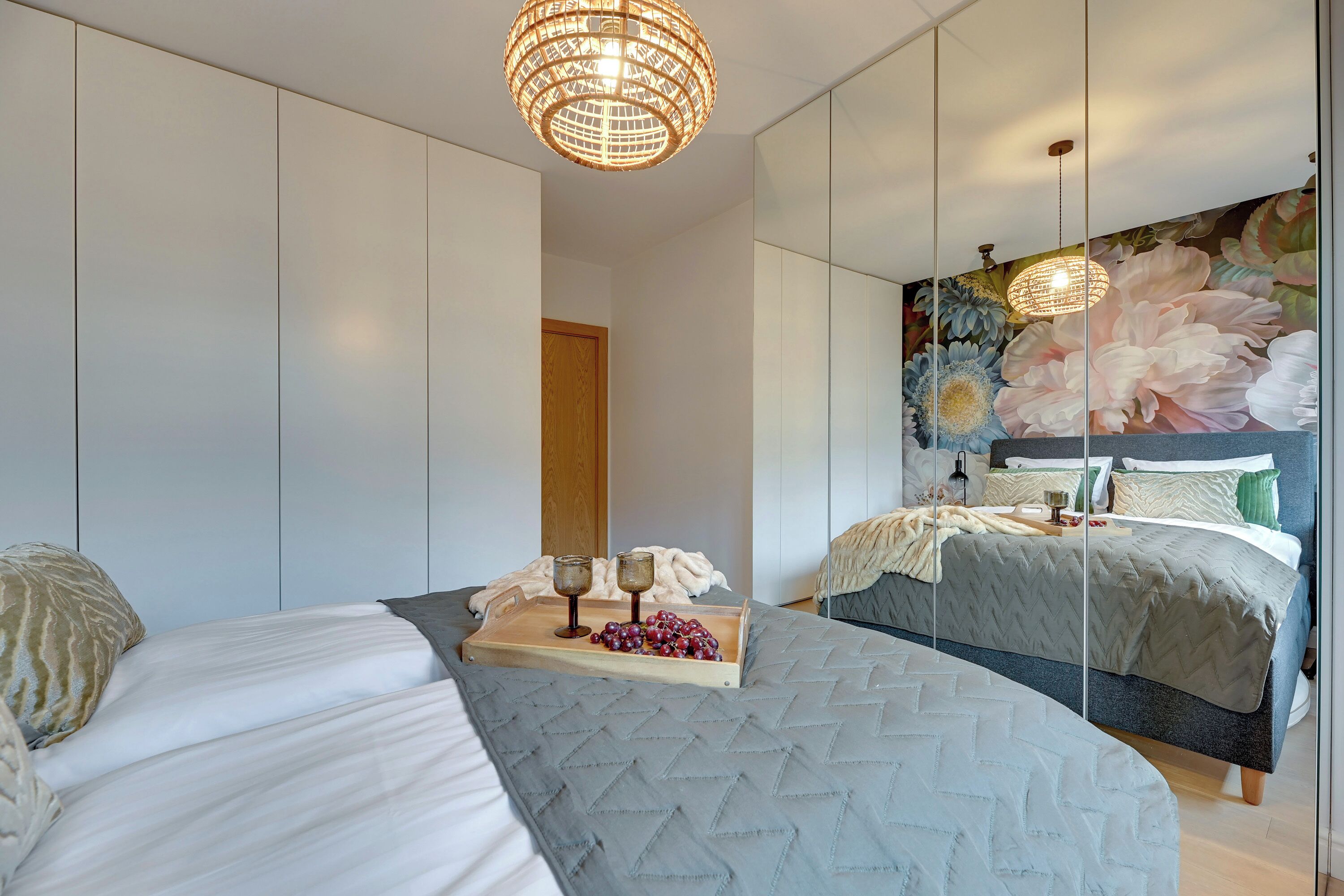 A modern bedroom with a large bed, a wooden bedside table with glasses and grapes, a mirror, and a floral wall mural.