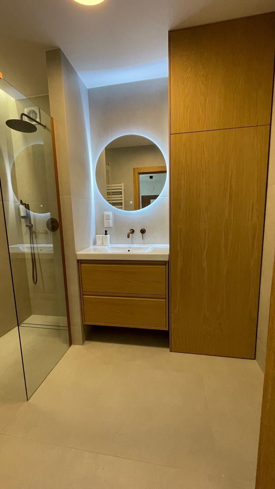 A modern bathroom with a glass shower enclosure, a round mirror, and wooden cabinetry.