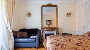 A room with a black leather sofa, a patterned bedspread, a large ornate mirror, and a fireplace with a decorative item on the mantel. - Parisian Opulence – Luxury Art Deco Apartment in Saint-Germain-des-Prés (Paris)