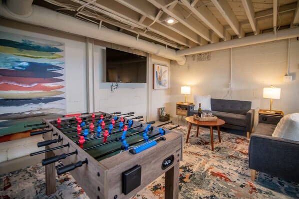 A room with a foosball table, a sofa, a lamp, and a painting on the wall.