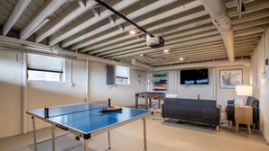Ein Zimmer mit Tischtennisplatte, einer Couch, einem Fernseher und einem an der Decke befestigten Projektor.