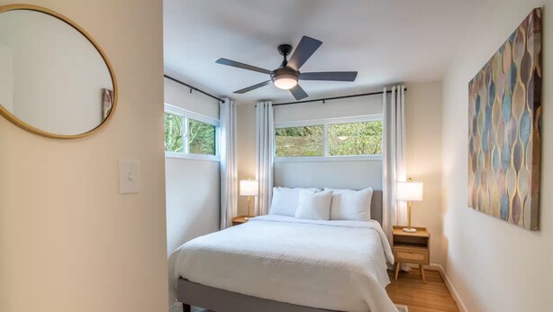 Ein Schlafzimmer mit einem Bett, Deckenventilator, zwei Nachttischlampen, einem Fenster und einem Bild an der Wand.