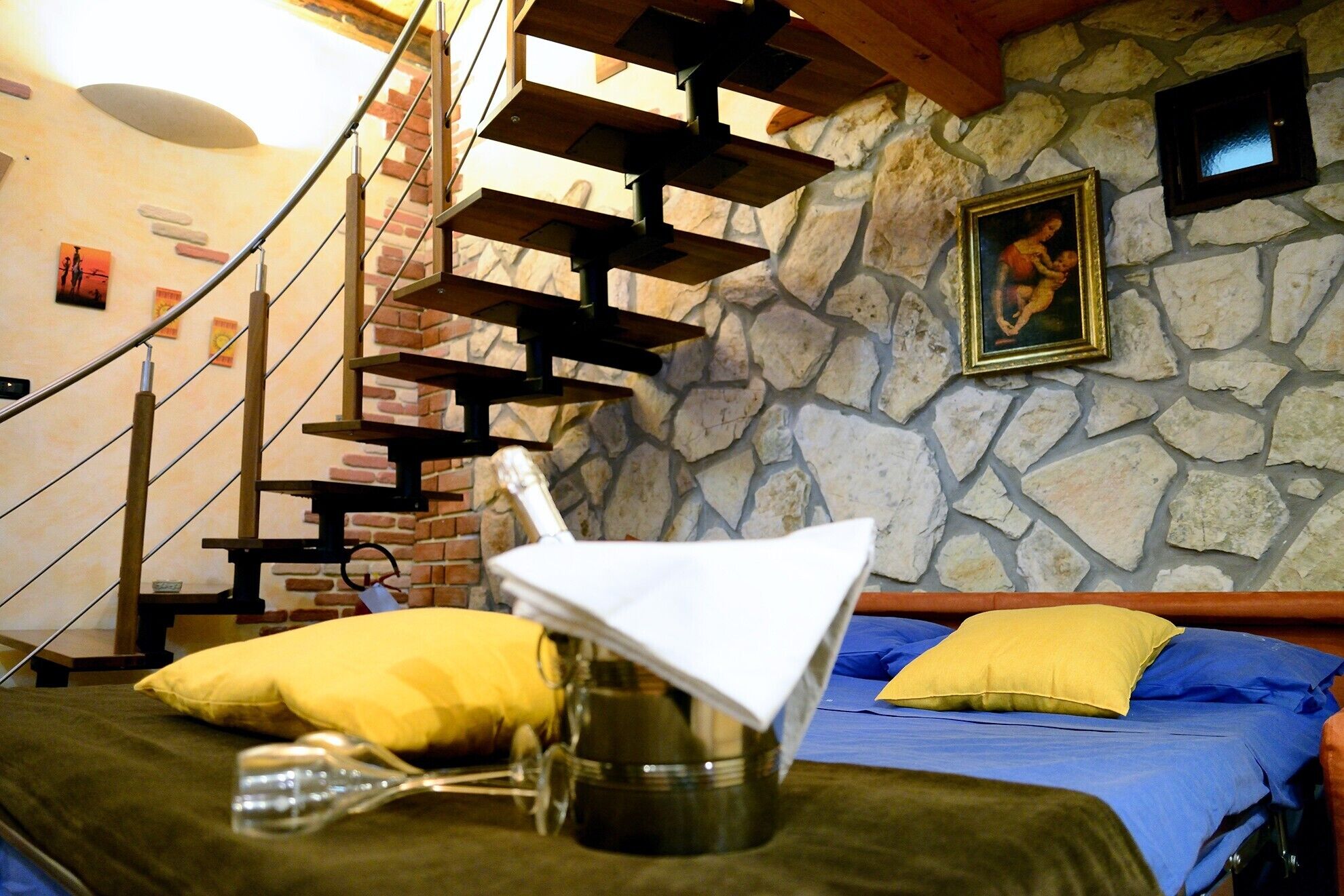 A room with a stone wall, a bed with blue and yellow bedding, a wooden staircase, and a framed picture on the wall.