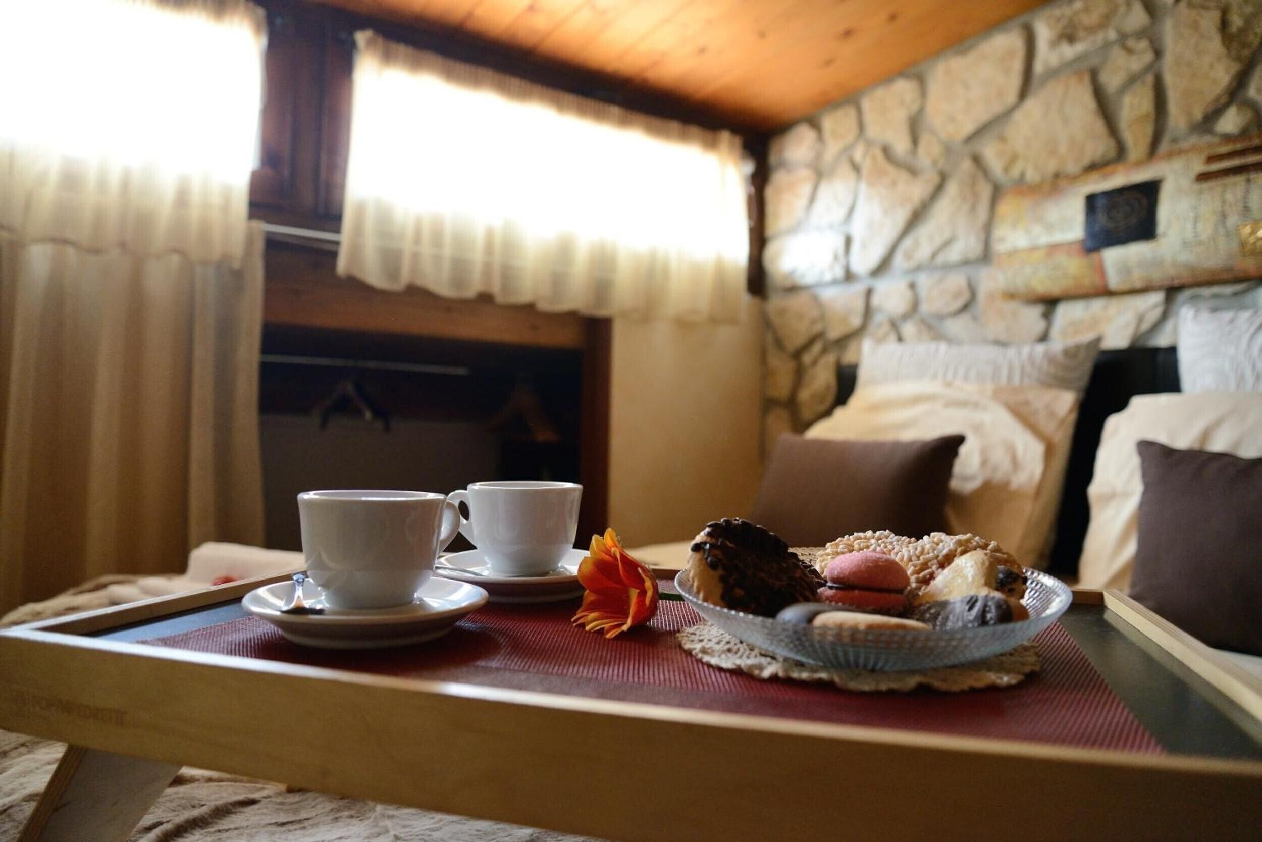 Un safata de matí amb dues tasses, una flor i pastissos sobre una taula en una habitació de parets de pedra i sostre de fusta.