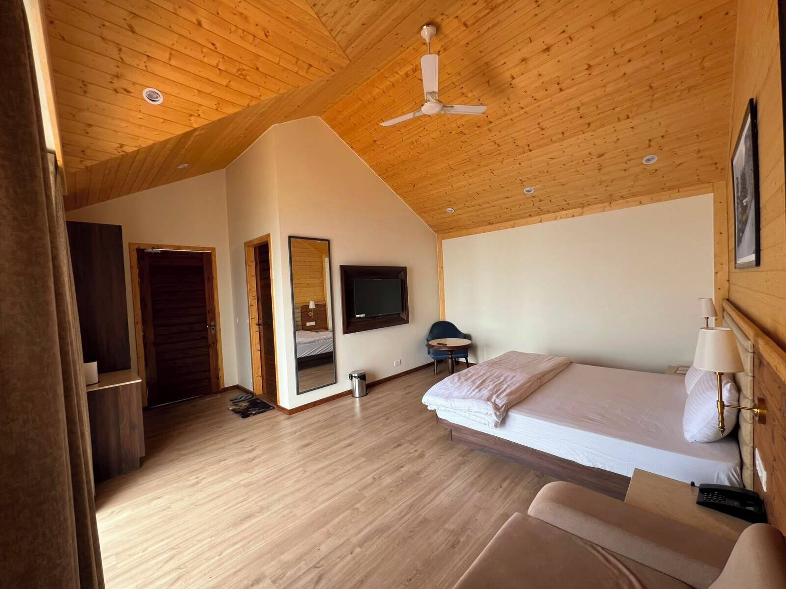A room with a sloped ceiling, wooden flooring, a bed, a chair, a desk, a television, and a mirror.