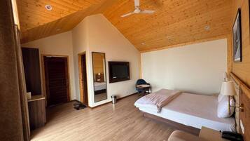 A room with a sloped ceiling, wooden flooring, a bed, a chair, a desk, a television, and a mirror.