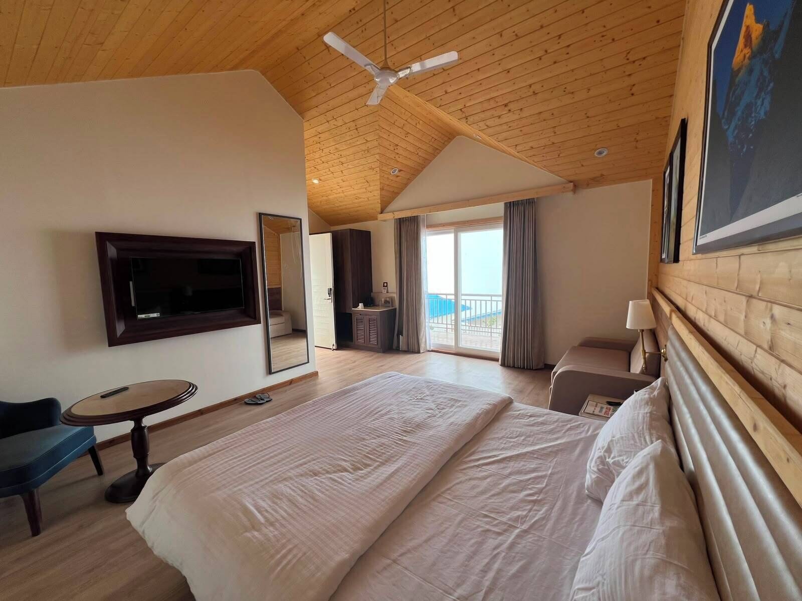 A hotel room with a large bed, a TV mounted on the wall, a desk, and a view of the outside.