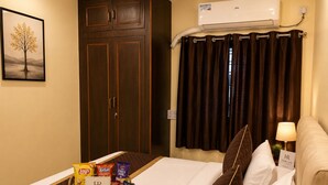 Quarto de hotel com uma cama, mesa de cabeceira com abajur, guarda-roupa de madeira escura, quadro na parede e janela com cortinas.