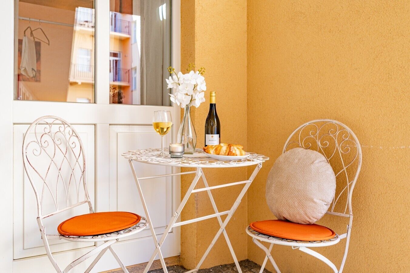 A small outdoor seating area with two white metal chairs with orange cushions, a round metal table, a bottle of wine, a glass of white wine, and a vase with white flowers.