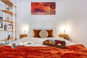Ein ordentlich bezogenes Bett mit roter Decke, ein Nachttisch mit Lampe, ein Bücherregal und ein Bild an der Wand.