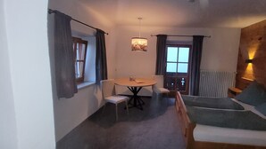 Una habitación con una mesa redonda, dos sillas, un sofá con cojines, una ventana con cortinas, un radiador y una lámpara fijada a la pared.