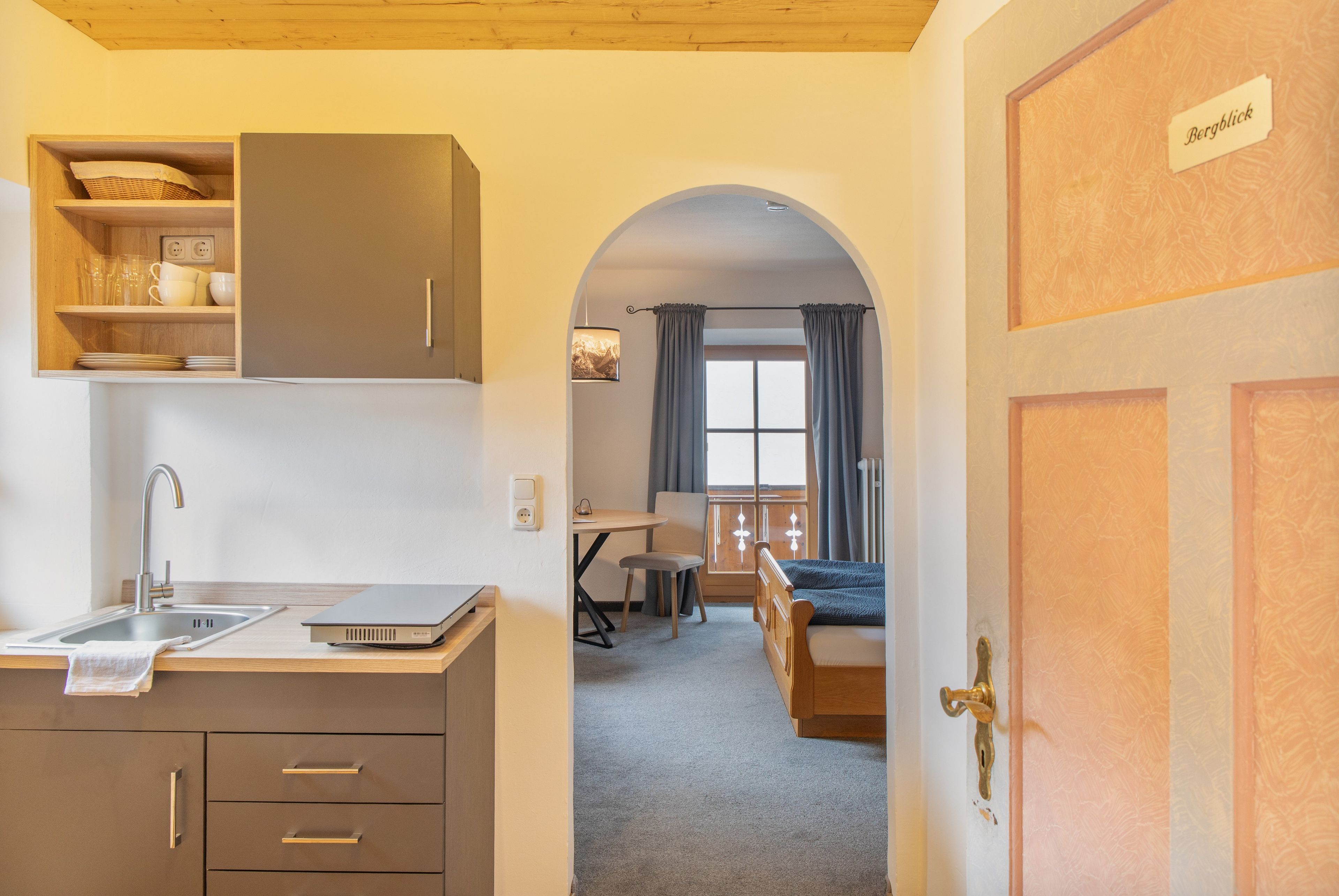 Cocina compacta con lavabo, armarios y encimera. Hay una zona de comedor con una mesa y sillas, y un dormitorio con cama y una puerta con la etiqueta 'Bergblick'.