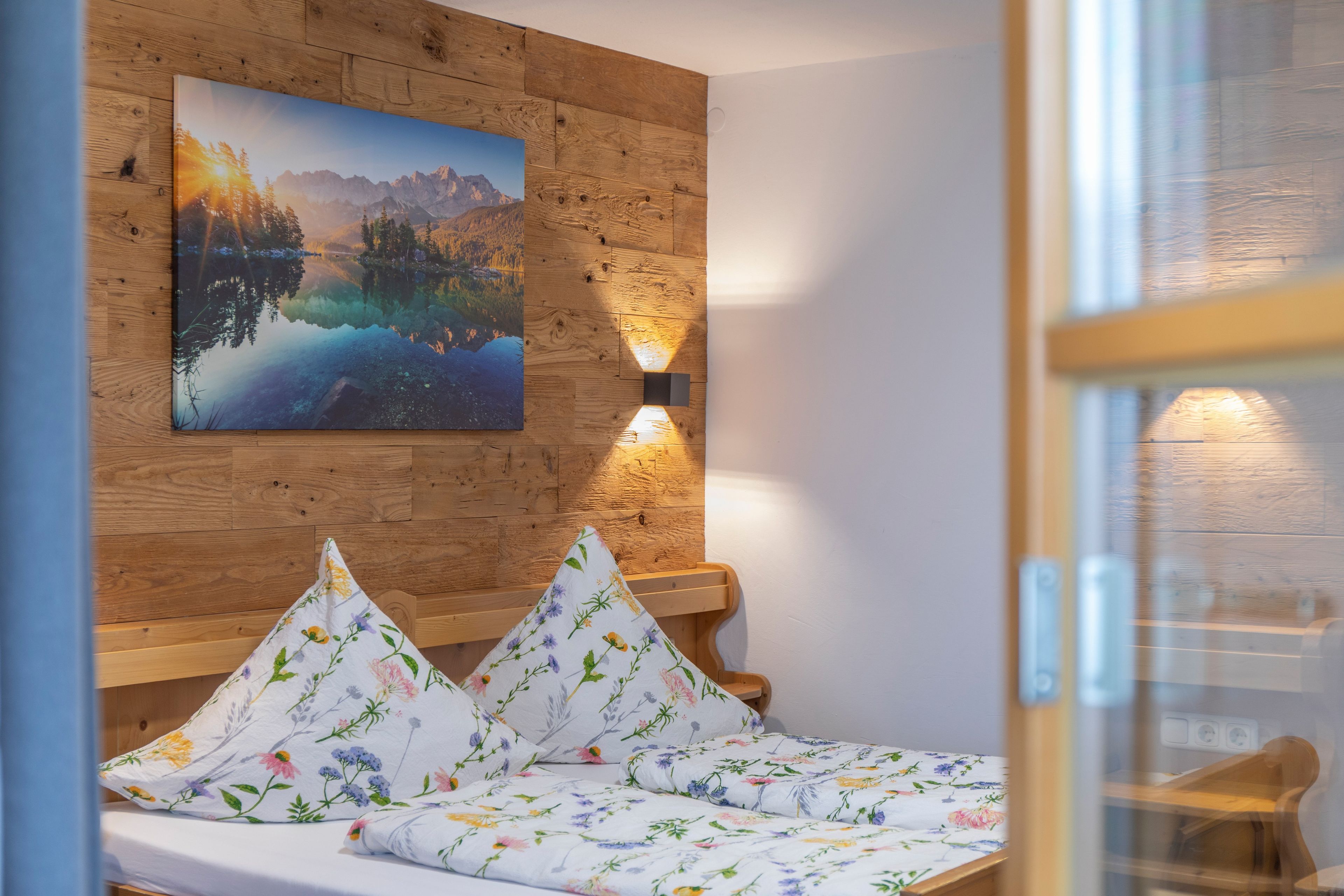 A room with a wooden wall, a bed with floral bedding, a painting of a mountain landscape, and a wall-mounted lamp.
