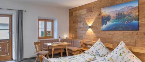 Una habitación con pared de madera, un cuadro de un paisaje montañoso, una cama con ropa de cama floral, una mesa con sillas y una ventana con cortinas.