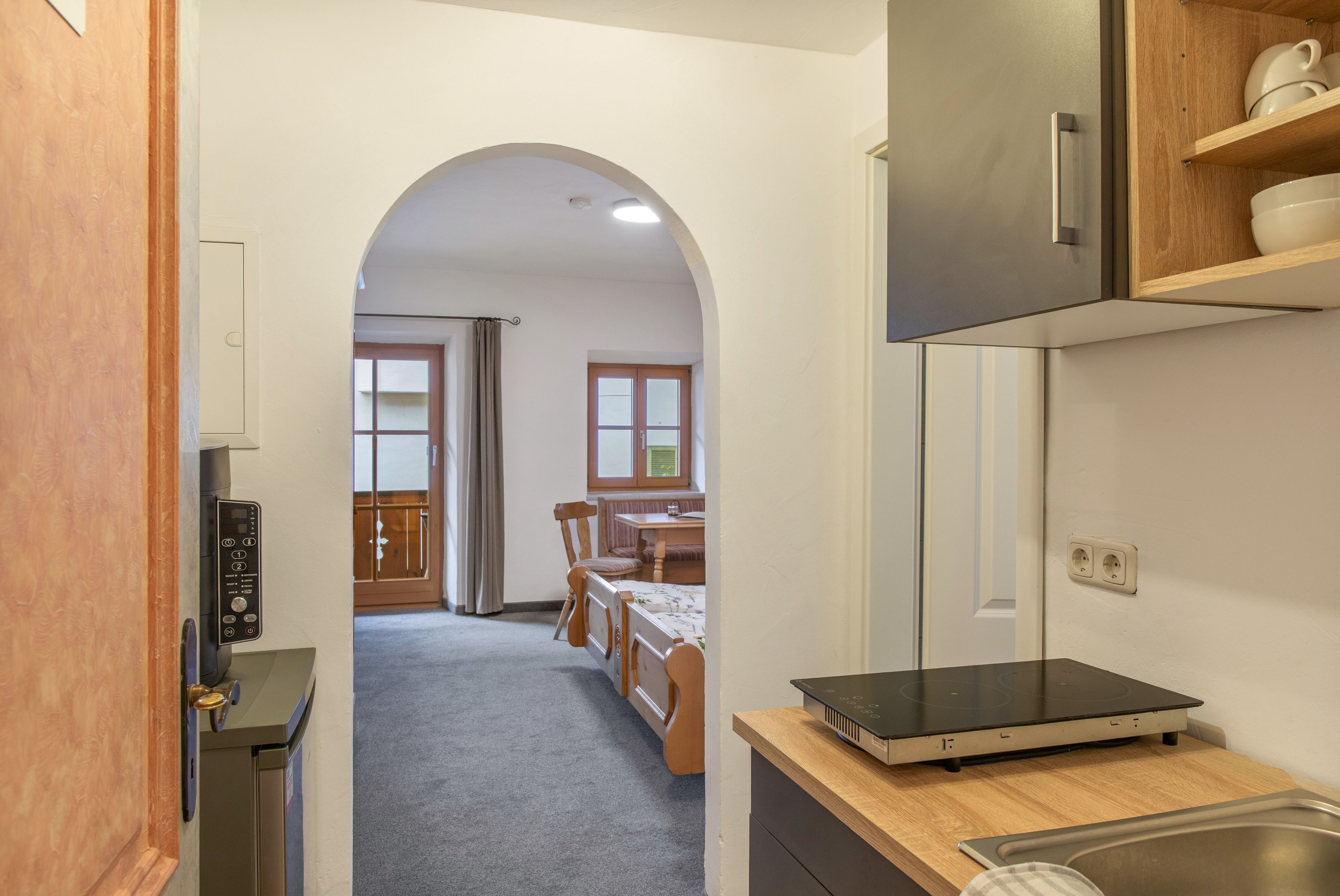 A narrow hallway leading to a dining area with a wooden table and chairs, a kitchen with a built-in oven, and a door with a control panel.
