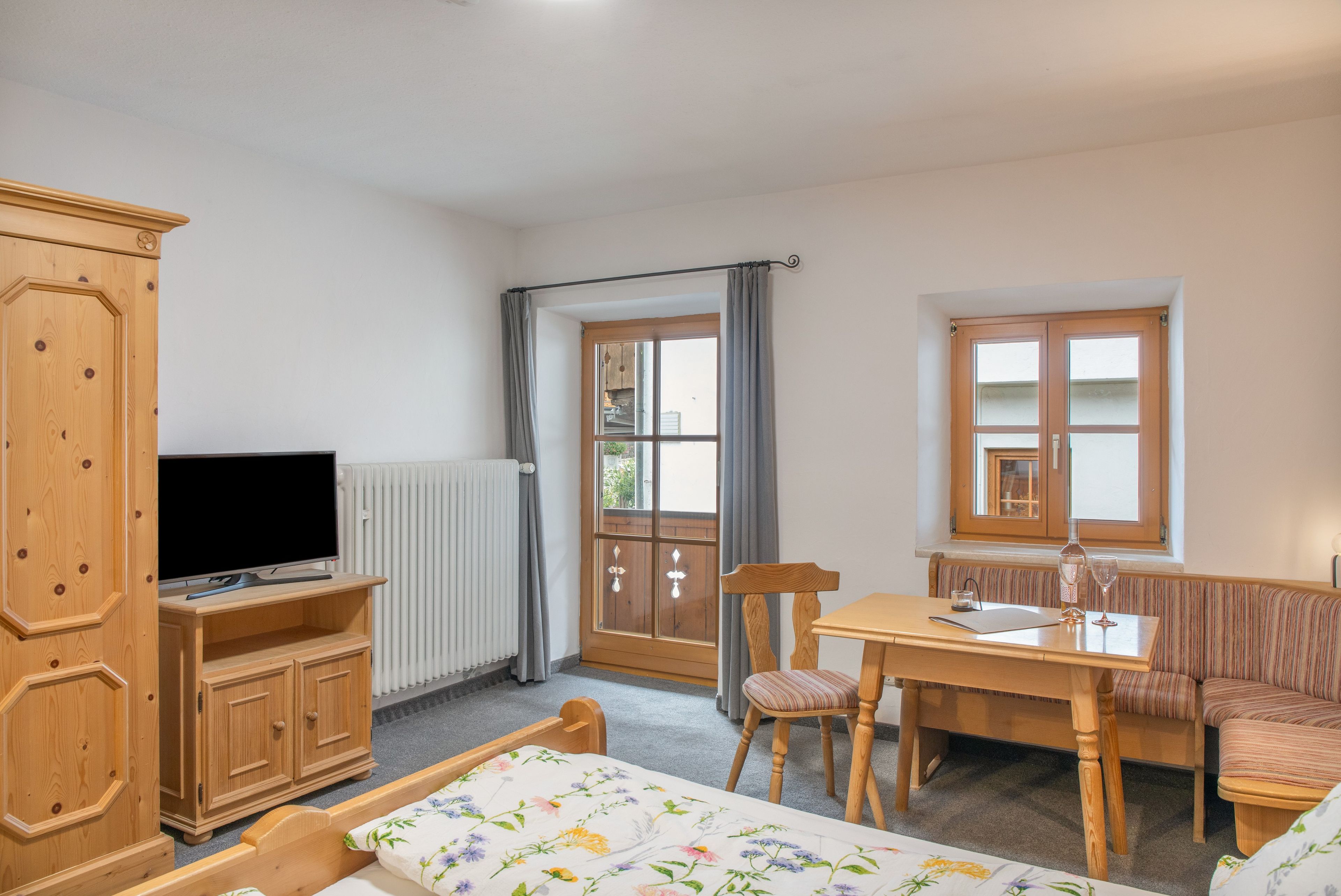 Quarto com cama, estante de TV de madeira, mesa e cadeiras, janela e porta.