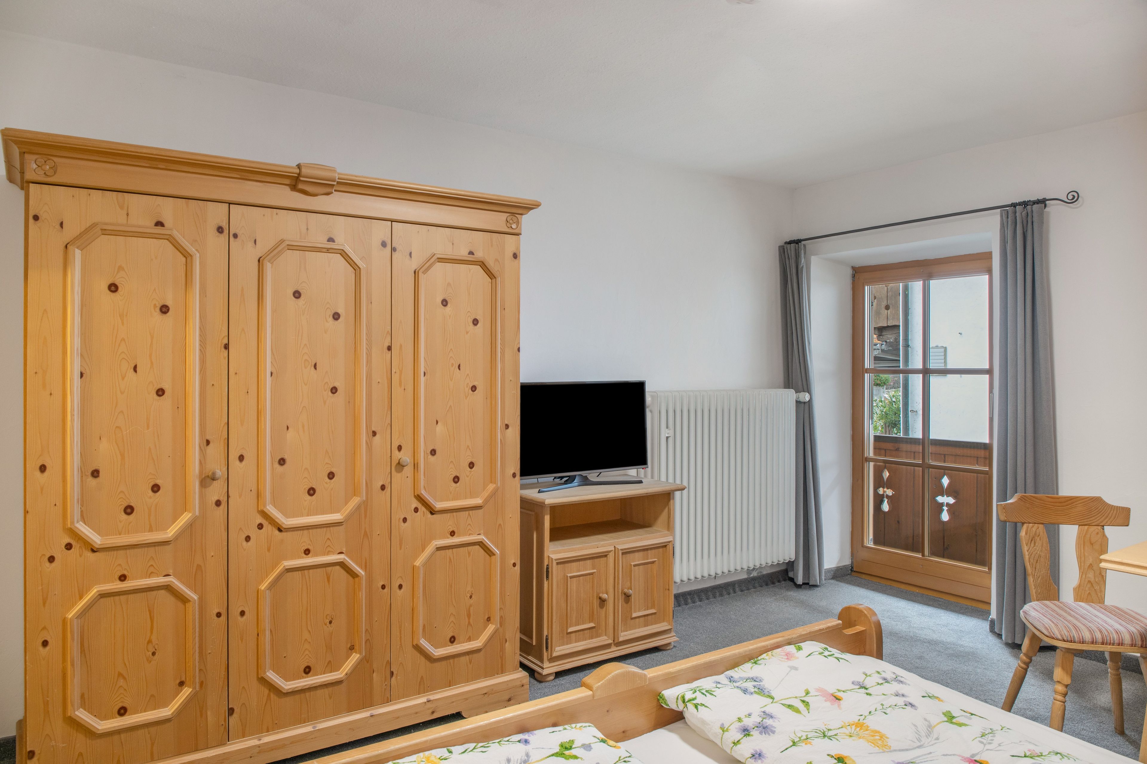 Ein Schlafzimmer mit Holzschrank, Fernseher auf einem Standfuß, Bett mit Blumenmuster-Bettwäsche, Stuhl und Fenster mit Vorhängen.