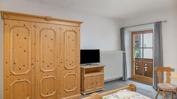 Ein Schlafzimmer mit Holzschrank, Fernseher auf einem Standfuß, Bett mit Blumenmuster-Bettwäsche, Stuhl und Fenster mit Vorhängen.