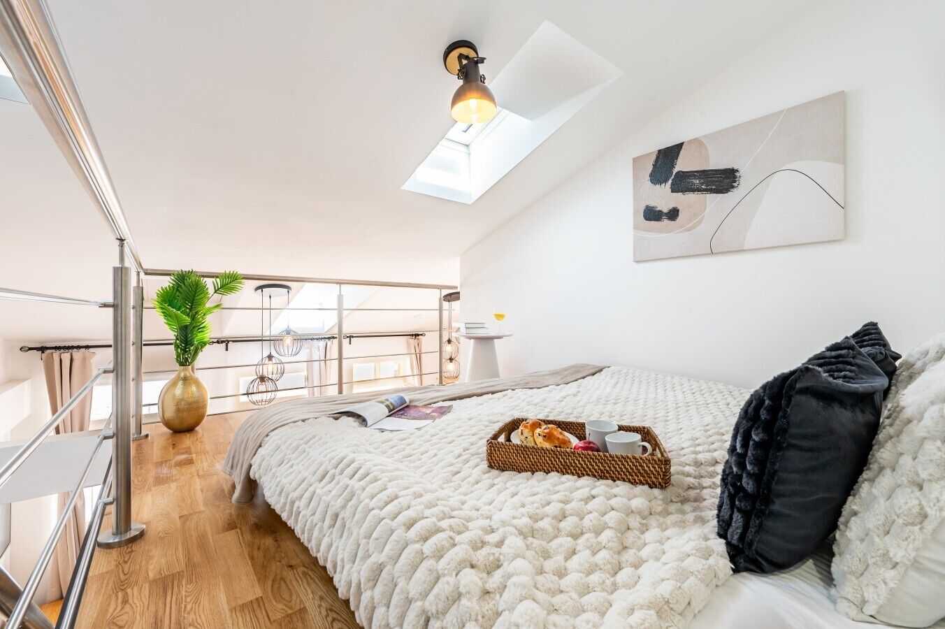Ein modernes Schlafzimmer mit Dachfenster, einem Bett mit weißer, strukturierter Decke, einem Nachttisch mit einer Platte mit Brot und Tassen sowie Blick auf einen Balkon mit Pflanzen und Lichterketten.