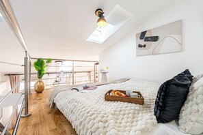 A modern bedroom with a skylight, a bed with a white textured blanket, a bedside table with a tray of bread and cups, and a view of a balcony with plants and hanging lights. - New Town City Loft with Free Parking (Prague)