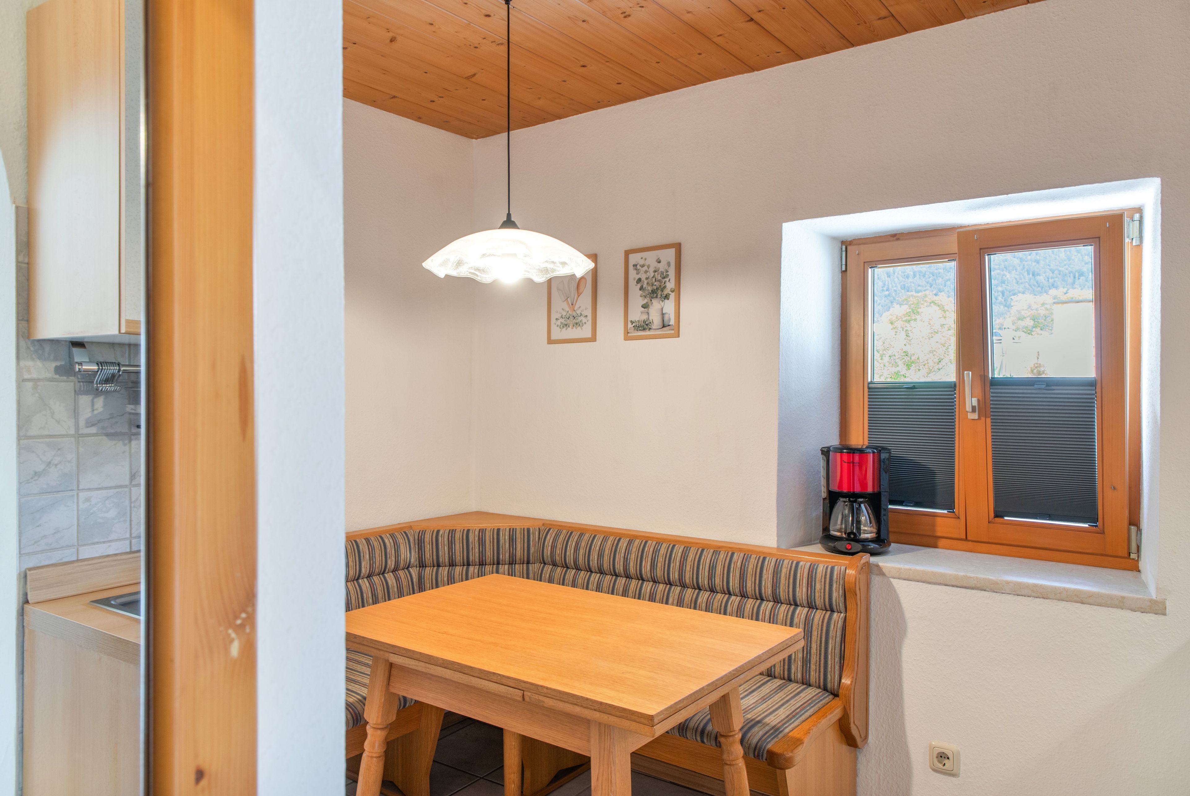 Ein Essbereich mit einem Holztisch, einer Bank, einer Pendelleuchte und einem Fenster mit Jalousien.