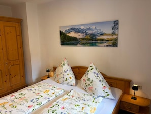 A bedroom with a bed, two pillows, a nightstand, a lamp, a wooden wardrobe, and a painting of a mountain landscape on the wall.