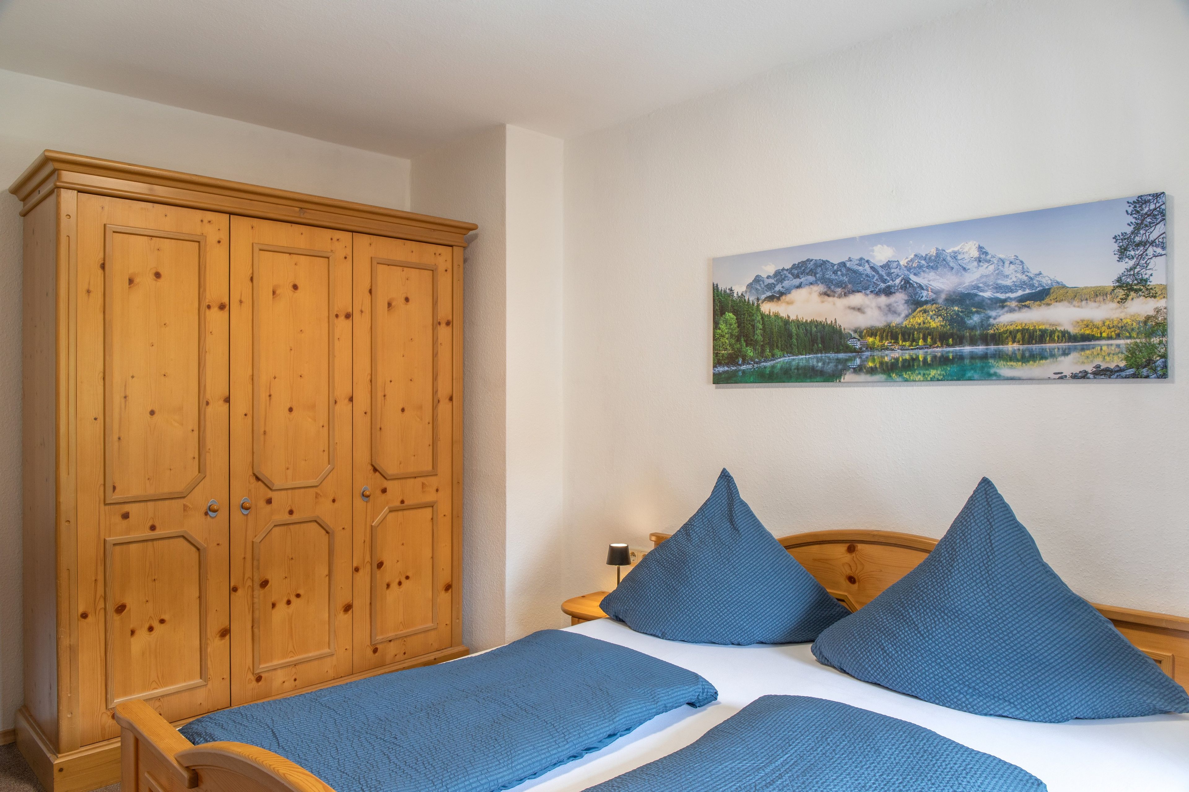 Un dormitorio con un armario de madera, una cama con ropa de cama azul y un cuadro de un paisaje montañoso en la pared.